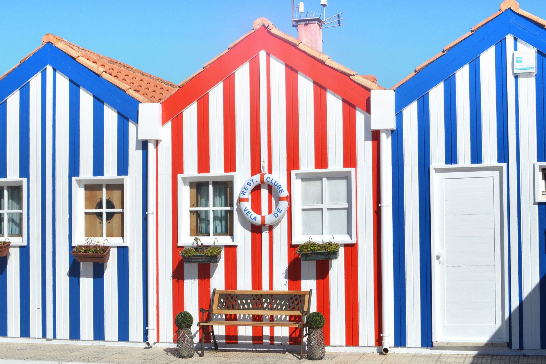 Brightly painted striped beach houses with a bench under sunny skies